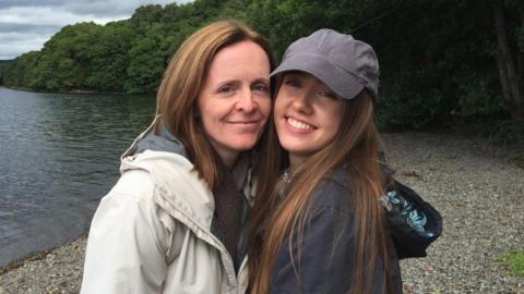 Beth, her mum, and Emily smile for the photo along a lake. Emily is wearing a grey cap and long brown hair whilst Beth has short gingery hair.