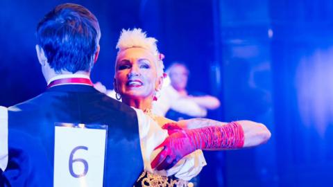 A female ballroom dancer with white blonde cropped hair, dancing with a man who has his back to the camera and a number six pinned on his waistcoat. The woman is wearing a long red glove and has red flowers in her hair.