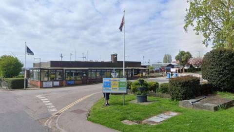 A google maps image of a security building outside HMP Standford Hill with a sign, flags waving and a grassy verge visible from the road