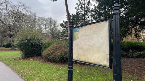 A sign in People's Park mounted on black metal legs which has been damaged leaving only a small corner and an expanse of cracked white plastic
