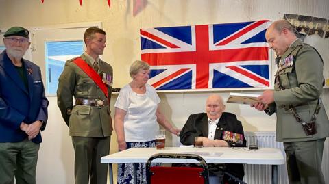 Henry Francis, a 100-year-old World War Two veteran, is seated at a table in front of a large Union Flag. He is surrounded by uniformed military personnel. One stands beside him, reading aloud a letter from Sir Gwyn Jenkins, First Sea Lord, as others look on.