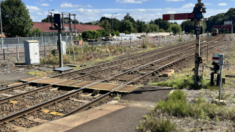 The barrow crossing at Hexham station. There is a panel under a section of the track for people to cross. There are signals and shrubbery on either side of the track and a building with a red roof to the left. A train looks to be approaching the station in the distance.