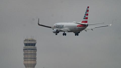 American Airlines plane seen from behind, flying through the air, with an air traffic control tower in the distance