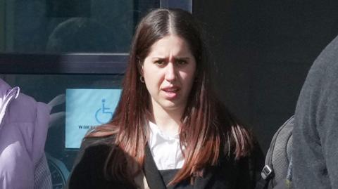 A woman with dark hair, wearing a black suit and white shirt. She is standing outside a court. 