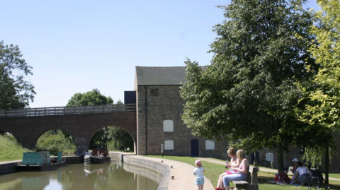 A large brick building and a bridge over a canal with a family seated on a bench in the foreground