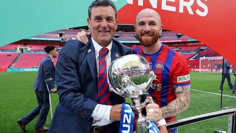 Then Aldershot Town manager Tommy Widdrington in blue suit and red and blue striped tie, with empty Wembley behind, holds FA Trophy