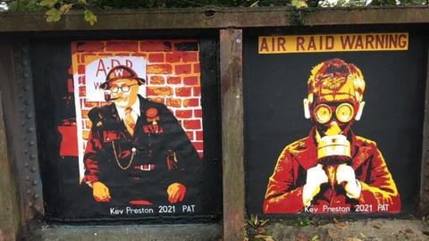 Two murals on the side of a bridge. On the left is a mural of an air raid warden wearing a helmet and uniform and smoking a pipe. A red brick wall with 'ARP' is behind him. On the right is a mural of a young boy wearing a gas mask with the words 'air raid warning' above him. Both paintings have been made using red, yellow and black. They both have the words 'Kev Preston 2021 PAT' at the bottom.