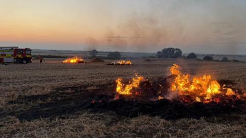 Bales in a field are on fire. The image is taken at dusk and the sunset can be seen through smoke in the distance. To the left is a fire engine and a firefighter is working close to it, to put out the flames from one of the bale sites.
