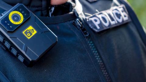 A close up of a police officer in uniform. They wear a black zipped up vest, with a police badge and body worn camera on the shoulder.