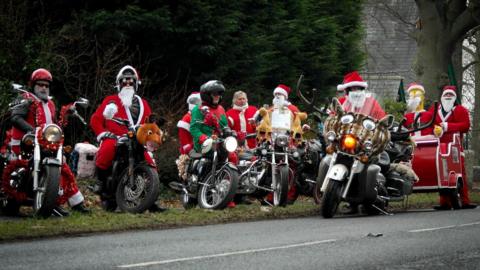 Five motorcyclists astride their vehicles on a grassy verge; four are dressed in Santa costumes, the other is in an elf outfit. All are adorned with tinsel and other decorations. The motorcycle on the right has a reindeer head on the front and is pulling a red sleigh, behind which stands another man in a Santa costume. Two of the other bikes also have pillion passengers both of whom are also in Santa outfits.