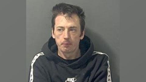 Tony McDonald in a police mugshot. The image is a bit out of focus. It shows him wearing a black and white Nike sweatshirt. He has dark hair, short at the sides and longer on top. He is looking over to his right.