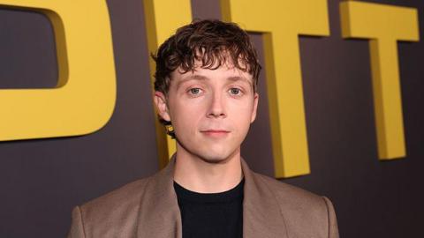 Gerran Howell, who wears a black jumper, beige suit jacket and has short brown hair