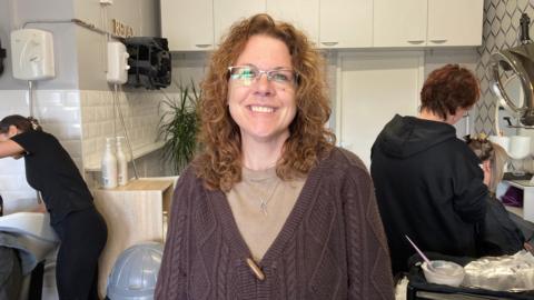 A woman standing in a hairdressers, wearing a brown jumper and a light brown top.