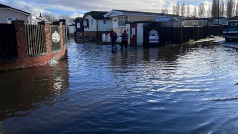 A flooded home park with two firefighters in waders walking through the floodwater.