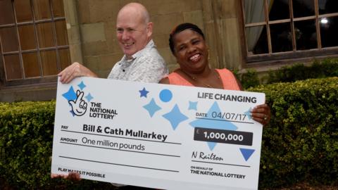 A couple smiling while holding a large blue and white cheque that reads 
"Bill and Cath Mullarkey, one million pounds."