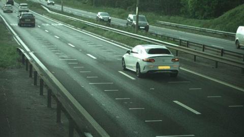 A speed camera image showing cars driving on a dual carriageway. The image is focused on a white Mercedes driving in the right-hand lane.