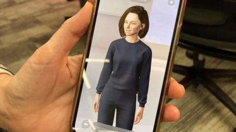 Sgrin ffon yn dangos chatbot AI sy'n gwisgo siwmper a throwsus glas tywyll a sydd a gwallt brown