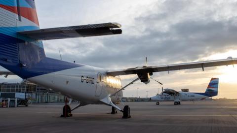 Two Skybus aircraft outside Guernsey Airport's main terminal building. The sun appears to be setting in the distance on a cloudy day.