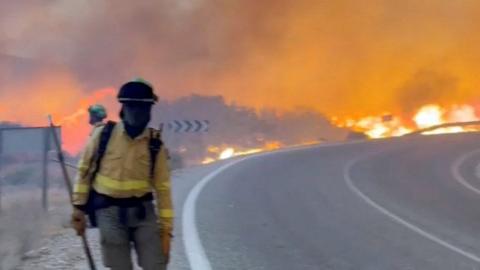 A masked emergency worker stands next to a road in Almería, Spain as a wildfire rages behind them