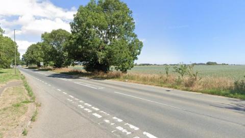 The view from a junction along a stretch of rural road with a worn-looking surface and faded white markings. Green trees stand on both sides under a blue sky, while fields with green crops stretch away to the right.