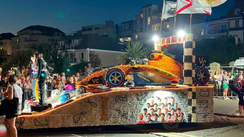 An illuminated Jersey Battle of Flowers parade float shaped like a Formula One racing car travels along a street at night. Spectators line both sides of the road behind barriers, while flags fly above the float and buildings are visible in the background.