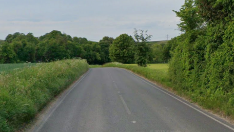 The A417 Wantage Road, near Streatley. It is a narrow, rural road with grass and bushes on either side.