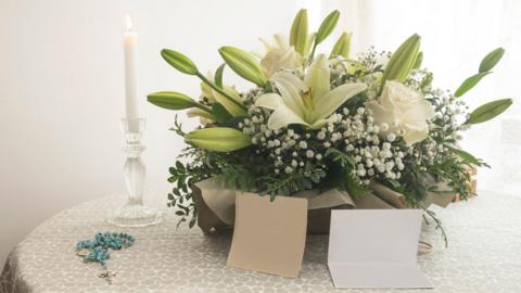 A white bouquet of flowers laid on a table with letters and a set of rosary beads. There is also a white candle on the table top, the scene is typical of a funeral service. 