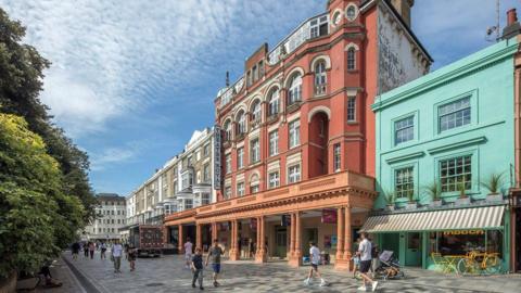 A large red-brick theatre building with cream detailing and arched windows stands on a pedestrian street, with people walking past and colourful shop fronts beside it under a bright sky.