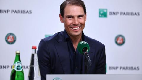 Rafa Nadal sits behind a microphone and smiles