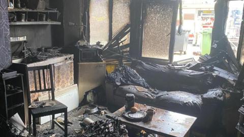 A room shows partially charred furniture and walls and objects like chairs, a stove, wallpaper and a other furniture covered in black soot. The windows are also blackened and panes of glass are missing. There are discarded bottles, plates and cups on the table and floor.