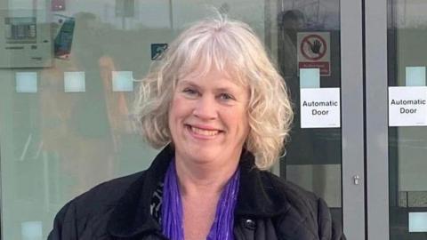 A woman with white, wavy hair who is smiling facing the camera. She is wearing a dark blue coat, blue scarf and black and white top. There are glass automatic doors behind her.