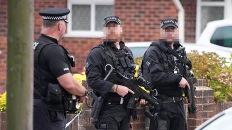 Three armed police at the scene of an incident in Lindum Road, Worthing. Two of the officers' faces are blurred out. All three are wearing black uniform and police caps. Two guns are visible in the image.
