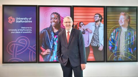 A man with short grey hair and a grey beard stands with one hand in his pocket and smiles. He is wearing a navy blue suit, white shirt and red tie. Behind him are some advertising screens with students from the University of Staffordshire.