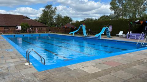 A view of the pool. It has a bright blue surround and the water is crystal clear. Two slides go into the pool on the side and there are steps at each corner.