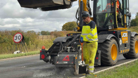 A large yellow JCB vehicle on a road. A workman in high-vis stands next to it.