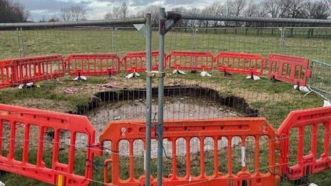 The large, round sinkhole is full of mud and water. It is surround by low plastic red fencing, with taller metal fences around it.  