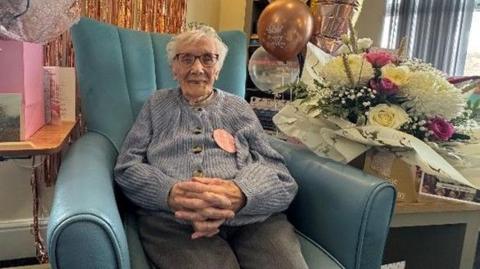 An elderly woman sat in a big blue arm chair in a blue cardigan smiling, with gifts, flowers and rose gold, pink and clear balloons. 