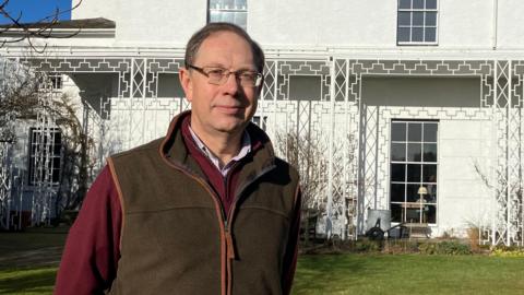 Richard Jebb pictured standing in front of the Lyth, a large white house. He is wearing a dark red fleece, with a brown gilet over the top, and is smiling slightly. The sky is blue and there is lots of strong sunshine. 