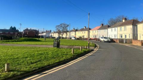 A road bend next to a children's park with a footpath running between it. On the other side of the road there's a row of attached houses, a couple of them have white cars parked in front of them. It's a sunny bright day with a clear blue sky. 
