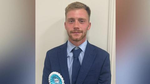 Reform UK councillor for Harraby South ward Josh Kirkwood. He is wearing a blue suit and tie and pale blue and white rosette.