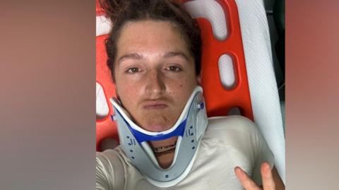 Matilda Pine wears a neck brace and lies on a hospital bed. She is holding her hand up and doing the peace sign with two fingers.
