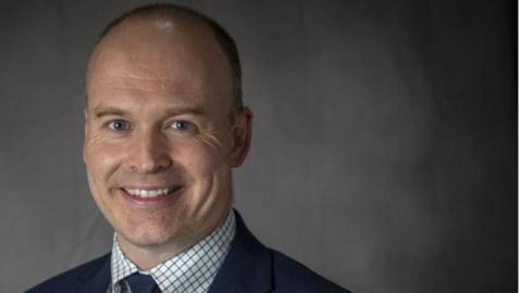 Head and shoulders shot of Andrew Crookham. He is wearing a blue suit, white and grey shirt and a blue tie. He has short hair and is smiling at the camera. He is standing in front of a grey backdrop.