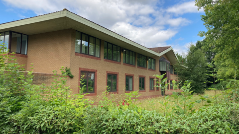 A two-storey light coloured brick building with windows on both floors. It is set back from the road, with bushes and trees in front