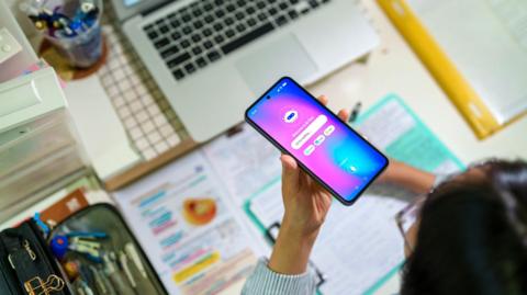 A young female student seen from above interacts with an AI chatbot on a smartphone while studying at a desk with a laptop, notes and stationery. The scene highlights modern learning and technology integration.