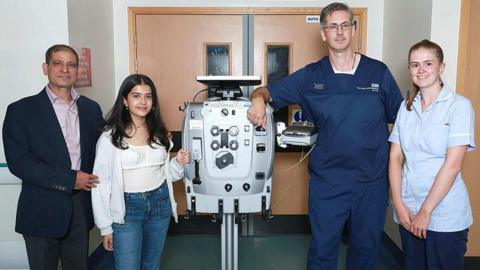 Two women and two men stand either side of a piece of hospital equipment in front of light brown double doors.