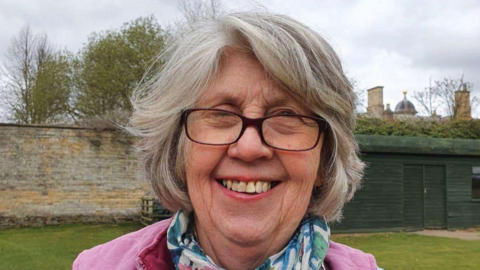 A woman standing outside on a grey day. She is smiling at the camera. She has short grey hair and is wearing glasses along with a pink jumper and a blue and green scarf. 