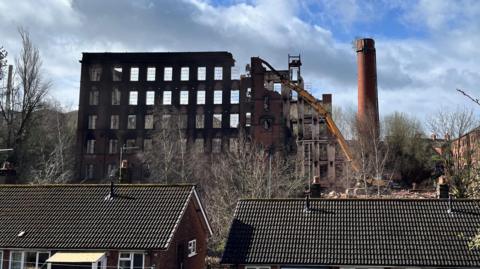 The shell of an old five or six-storey mill is being demolished by a large yellow vehicle. It is behind some trees and some houses.