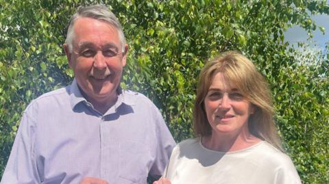 The Souttars in blue and white shirts standing side by side smiling in front of a tree