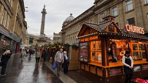 A number of wooden stalls in this year's Christmas Market, including a churros stall in the foreground on the right-hand side. Several people are walking along the street. The pavement is wet as it is raining. A number of ornate stone buildings can be seen on either side with Grey's Monument - a stone column - rising in the background.