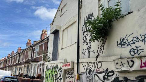An empty cream-rendered property with boarded windows is covered in graffiti with a bush growing from a windowsill. Next to it is a row of red brick terraced houses on a residential street.